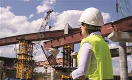 A female construction worker holding a laptop and wearing a yellow hi-vis vest and white hard had inspects a construction site.