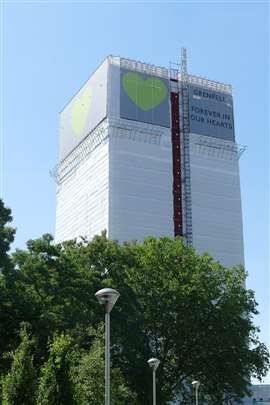 An image of Grenfell Tower after the 2017 fire, with a white and green hoarding erected around it reading "Grenfell: Forever in our hearts".