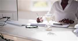 A woman sitting at an office desk taps on a calculator with an hourglass in front of her.