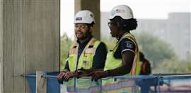 Two Clark Construction workers in discussion, wearing yellow hi-vis jackets and white hard hats.