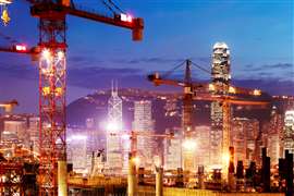 Construction cranes in Hong Kong at night with skyscrapers in the background.