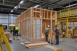 Construction workers assembling modular wooden home in factory with tools and scaffolding.