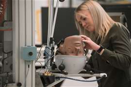 Una tecnica di laboratorio Mips tiene in mano la testa di un manichino per crash test, indossando un casco industriale bianco su un banco di prova.