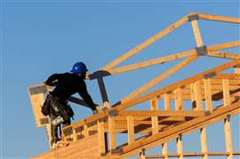 A construction worker assembles wooden trusses on the roof of a residential building. (Image: Adobe Stock)