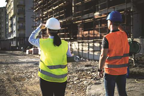 A female construction worker in a yellow hi-vis vest and white hard hat, and a male worker in an orange hi-vis vest and blue hard hat inspect a construction site.